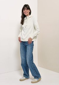 Junge Frau trägt einen weißen Hoodie, blaue Jeans und weiße Sneakers, steht und lächelt vor einem neutralen Hintergrund.