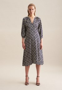 Granatowa sukienka z dekoltem w serek i rękawami 3/4 z bufkami, ozdobiona geometrycznym białym wzorem i w długości midi. Czarne sandały na obcasie.