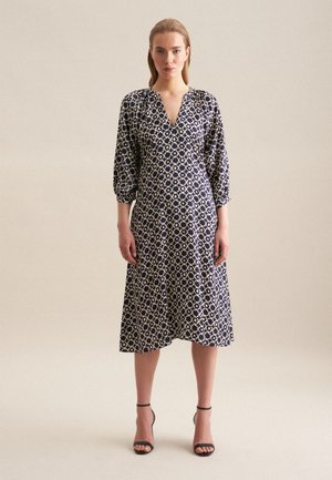 Donkerblauwe jurk met een V-hals en driekwart pofmouwen, met een geometrisch wit patroon en een middellange silhouet. Zwarte hakken sandalen.