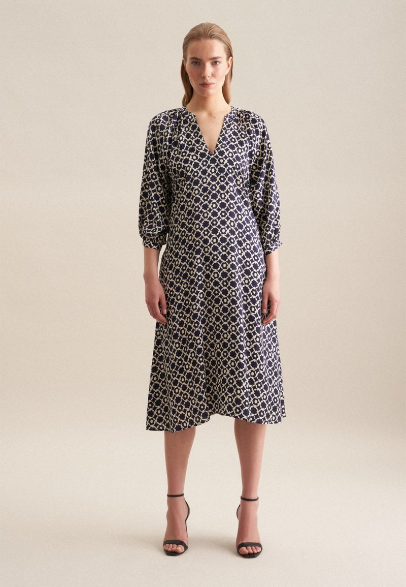 Granatowa sukienka z dekoltem w serek i rękawami 3/4 z bufkami, ozdobiona geometrycznym białym wzorem i w długości midi. Czarne sandały na obcasie.