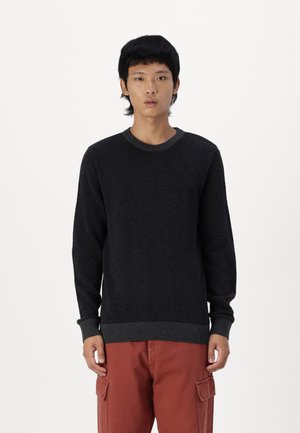 Pull en maille sombre avec une surface texturée, encolure arrondie et manches longues, avec des poignets et un ourlet côtelés. Porté avec un pantalon cargo orange.