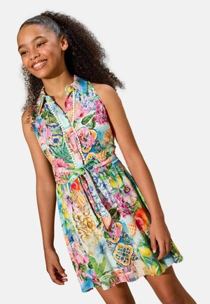 Mädchen mit lockigem Haar, das ein ärmelloses, kragenloses Kleid mit bunten Blumen- und Muschelmuster trägt, lächelt und steht vor einem einfarbigen Hintergrund.