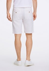 White linen shorts with a straight cut. Features two back pockets with circular buttons. Worn with white sneakers.
