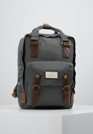 Sac à dos en tissu gris avec des sangles en cuir marron et des boucles en laiton, une poche avant et une poignée supérieure sur un fond blanc uni.