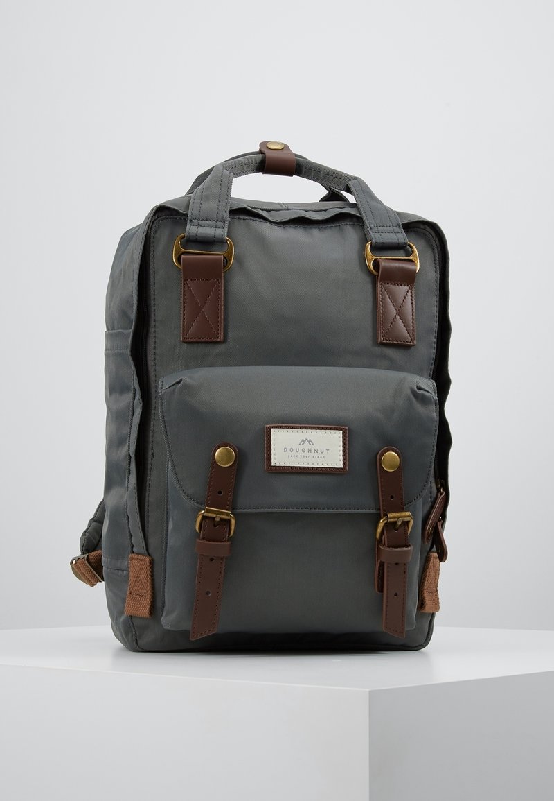 Sac à dos en tissu gris avec des sangles en cuir marron et des boucles en laiton, une poche avant et une poignée supérieure sur un fond blanc uni.