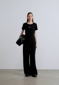 Črna bombažna majica z majhnim logotipom, kombinirana s črnimi širokimi hlačami in prevelikim črnim usnjenim plaščem, nošena s črnimi čevlji.