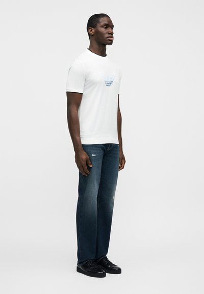 Άνδρας όρθιος που κοιτάει εμπρός, φοράει λευκό γραφικό μπλουζάκι, σκούρο μπλε τζιν με ελαφριά φθορά και μαύρα αθλητικά παπούτσια, μπροστά σε απλό φόντο.