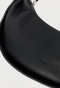 Μαύρη δερμάτινη τσάντα χειρός με λεία υφή και καμπυλωτό σχήμα. Διαθέτει το λογότυπο DKNY ανάγλυφο στην επιφάνεια. Ασημένια μεταλλικά στοιχεία ως διακοσμητικό.