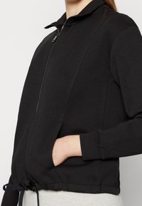 Zwarte zip-up jas van zacht materiaal, met een kraag, zijzakken en een trekkoord aan de onderkant voor aanpassing. Fitted stijl.
