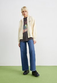Kremfarget zip-up hettegenser over en svart grafisk t-skjorte, kombinert med vide, blå denimjeans og svarte snøresko. Flatt grønt gulv som bakgrunn.