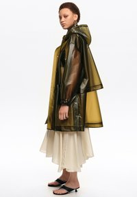 Jeune personne portant un imperméable translucide vert olive avec capuche, une jupe midi crème, des sandales noires à talons, se tenant de profil sur un fond blanc.