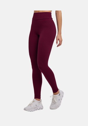 Frau trägt hoch taillierte, strukturierte, bordeauxrote Leggings und weiße Sportschuhe, steht vor einem schlichten hellen Hintergrund.
