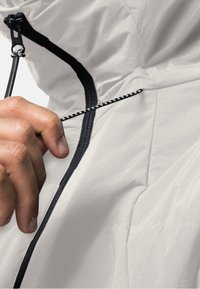 Main en train d'ajuster un cordon noir et blanc sur une veste claire avec une fermeture éclair noire visible et une texture de tissu.