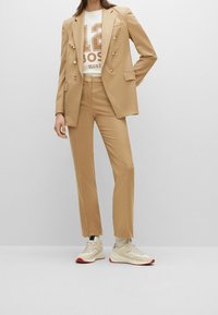 Setelan tailoring beige dengan jas double-breasted, celana slim matching, dan kaos grafis putih. Dipadukan dengan sepatu sneaker beige dengan aksen merah.