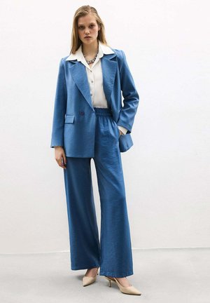 Ung kvinde iført blå bukser med vidde og matchende blazer over en hvid bluse, accessoriseret med halskæde og beige hæle.