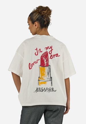 Göndör hajú nő, fehér, túlméretezett pólót visel, amely hátul egy nagy piros rúzsillusztrációt és szöveget ábrázol.