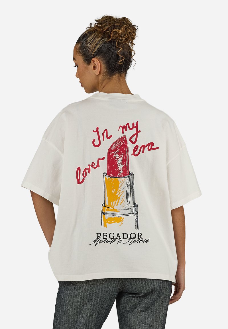 Γυναίκα με σγουρά μαλλιά που φοράει ένα λευκό φαρδύ t-shirt με μεγάλη εικονογράφηση κόκκινης κραγιόν και κείμενο στην πίσω πλευρά.