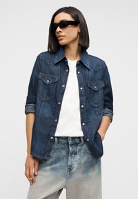 Γυναίκα που φοράει σκούρο τζιν μπουφάν πάνω από λευκό πουκάμισο, ανοιχτόχρωμο ξεβαμμένο τζιν και μαύρα γυαλιά ηλίου, με το ένα χέρι στην τσέπη, μπροστά σε απλό φόντο.