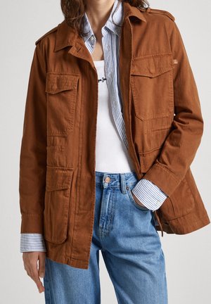 Personne portant une veste marron sur une chemise rayée bleu et blanc et un débardeur blanc, associée à un jean en denim bleu clair.