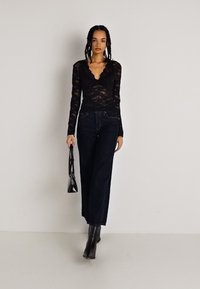 Zwarte kanten top met lange mouwen en een gebogen halslijn, gecombineerd met wijde donkerblauwe jeans en zwarte hakken, terwijl ze een zwarte handtas vasthoudt.
