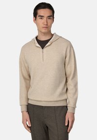 Sweatshirt beige avec une fermeture éclair quart de tour, capuche et poignets côtelés. Texture douce, coupe décontractée. Porté avec un sous-vêtement marron foncé et un pantalon gris.
