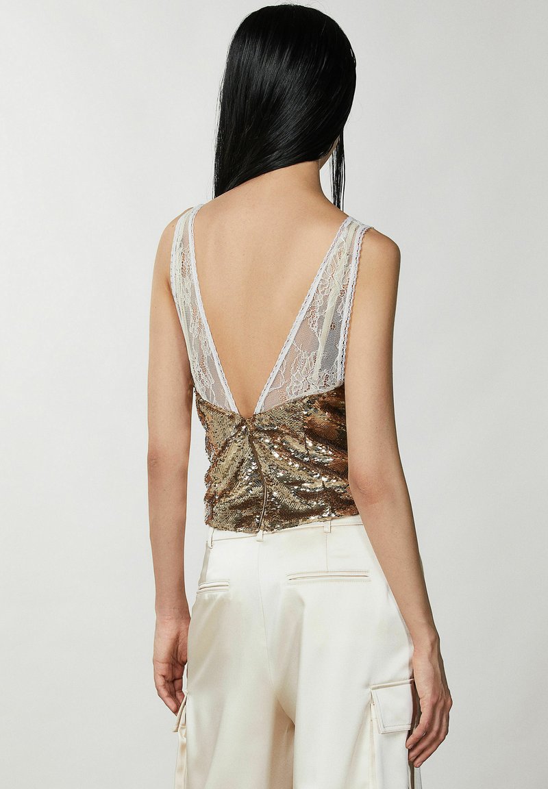 Złota bluzka z cekinami i koronkowym dekoltem w kształcie litery V z tyłu, z cienkimi ramiączkami i efektem marszczenia. Zestawiona z kremowymi, satynowymi spodniami cargo.
