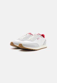 Weiße Sneakers mit grauen Akzenten, ausgestattet mit einer strukturierten Gummisohle, rotem Innenfutter und auffälligem Logo an der Seite. Klassisches Schnürdesign.