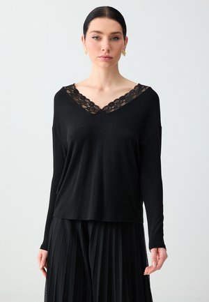 Woman with dark hair in a low bun wearing a black long-sleeve top with lace V-neckline and black pleated skirt, standing against a plain background.