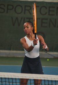 Giocatore di tennis con una canotta bianca e una gonna blu scuro, che tiene una racchetta da tennis arancione, pronto a colpire una palla su un campo con una rete.