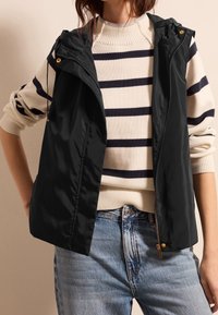 Black sleeveless vest with a smooth, shiny texture and a zip front. Worn over a cream sweater with navy horizontal stripes.