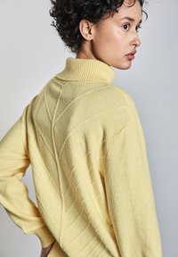 Gelber Strickpullover mit einem hohen gerippten Kragen, der auf der Rückseite strukturierte Chevron-Muster und gerippte Ärmelbündchen aufweist.