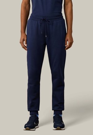Persona que lleva pantalones deportivos azul marino con cintura de cordón y zapatillas azul marino sobre un fondo neutro, de pie y recto.