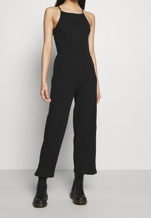 Sort ribbet jumpsuit med tynde stropper, tætsiddende overdel, brede ben og struktureret stof. Kombineret med sorte snørestøvler til anklen.
