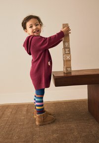 Kind in einer burgunderfarbigen Strickjacke mit Tasche, gestreiften Leggings und beigen Stiefeln, stapelt Holzklötze mit Buchstaben und Zahlen.