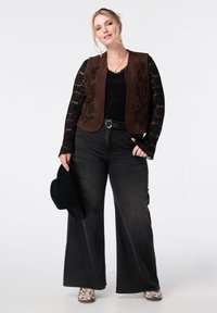 Vrouw die een zwarte kanten top draagt, een bruin geborduurd vest, zwarte wijde jeans, een zwarte hoed vasthoudt en met gespikkelde witte laarzen aan heeft.