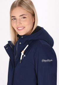 Marinblå jacka med huva, med dragkedja och tryckknappar. Vit snodd och detaljer i träpärlor. Varumärkeslogotyp på ärmen.