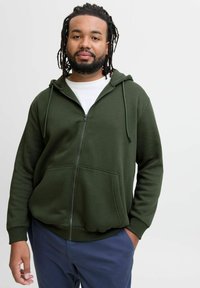 Olijfgroene zip-up hoodie met een trekkoordkap en voorzakken, gemaakt van zachte stof, gedragen over een wit T-shirt, gecombineerd met marineblauwe broek.