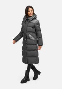 Manteau d'hiver long gris avec capuche, comportant des sections matelassées, une poche avant et une taille cintrée. Porté avec des bottes noires et un pantalon.