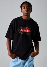Camiseta negra de manga corta con un gran logo rojo y dorado que dice "GRMY" en la parte delantera. El material parece suave y tiene un corte relajado.
