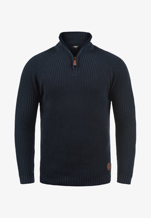 Sötétkék kötött pulóver magas gallérral és fényzippel, bordás mandzsettákkal és hemmel, valamint egy kis bőrpárkány-logóval az oldalon.
