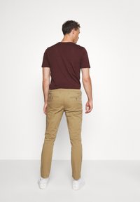 Bordeauxrode t-shirt gecombineerd met tan chino's. De chino's hebben achterzakken met knoopsluitingen. Het model draagt witte sneakers. Eenvoudig, goed aansluitend ontwerp.