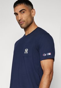 Jūras spēku kokvilnas T-krekls ar krūšu kabatu, izšūtu Ņujorkas Jankiju emblēmu un Champion un MLB logotipiem uz piedurknes.
