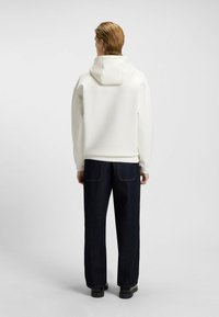 Hvid hættetrøje med en glat tekstur, parret med mørke jeans, der har synlige syninger og to baglommer.