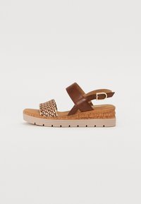 Brown leather sandal with woven strap, cork sole, adjustable buckle, and contoured footbed. Features a low wedge heel and textured details.