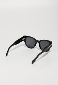 Zwarte cat-eye zonnebril met donkere lenzen, glanzende afwerking, slanke pootjes en een gouden accent op de veren, met gegraveerde merkinformatie.