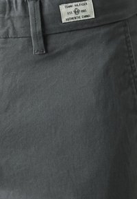 Pantalons chino en coton gris avec une texture lisse et une étiquette indiquant "Tommy Hilfiger Authentic Chino." Comprend une taille standard et des coutures.