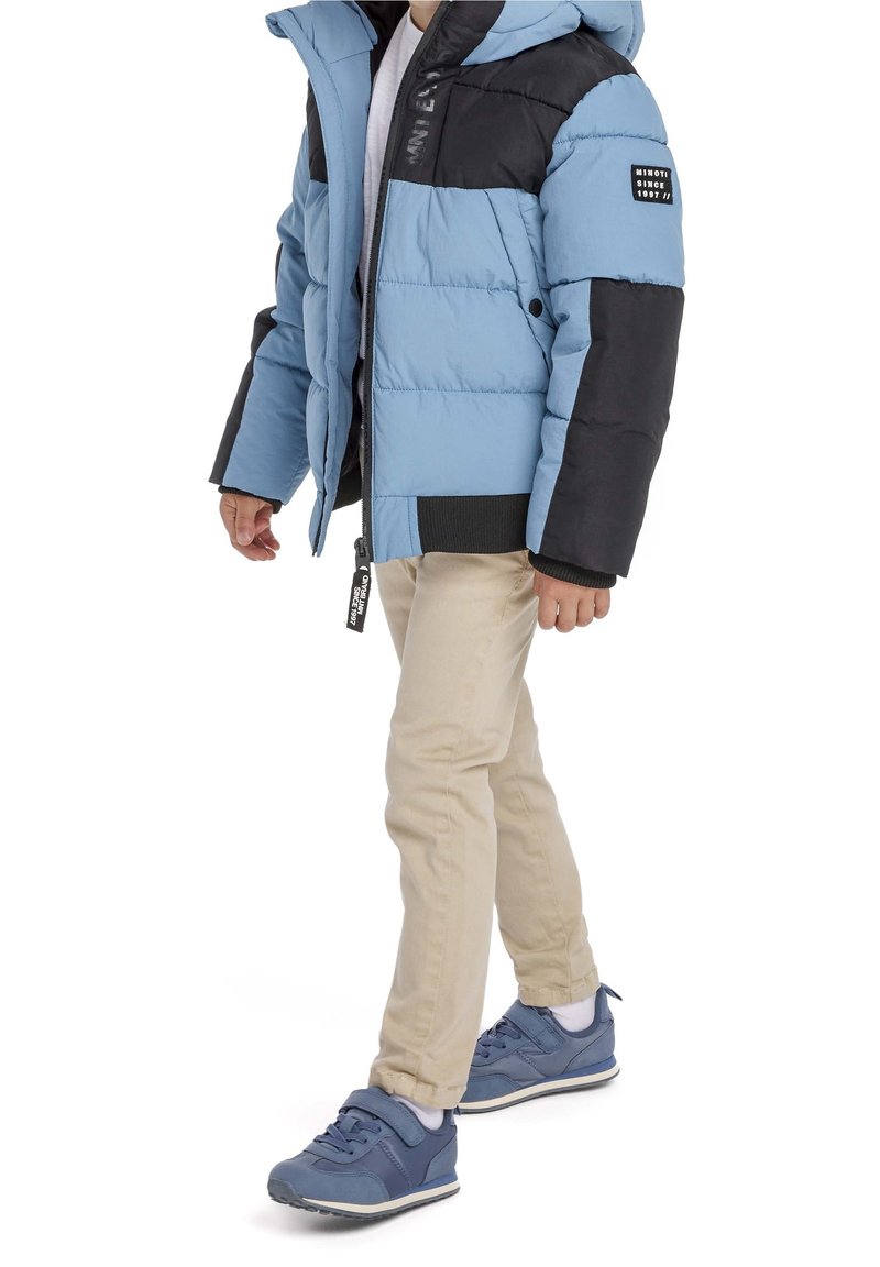 Enfant portant une veste matelassée bleu clair et noire, un pantalon beige, des chaussettes blanches et des baskets bleues, debout sur fond blanc.