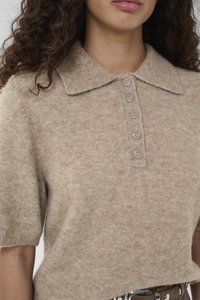 Frau trägt ein beiges, kurzärmeliges Strickoberteil mit weitem Kragen und fünf Knöpfen in der Mitte vorne.