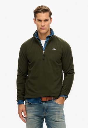 Green fleece pullover with a half-zip collar, worn over a denim shirt. Notable logo on the chest. Paired with blue jeans and a brown belt.