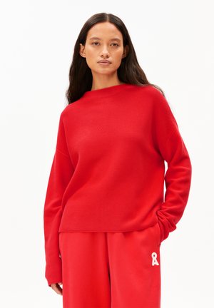 Roter Strickpullover mit hohem Kragen, überschnittenen Schultern und langen Ärmeln. Kombiniert mit passenden roten Jogginghosen, die ein weißes Band als Akzent haben.